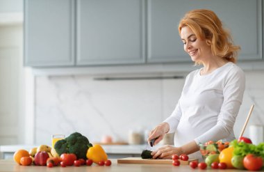 Hamile bir kadın şık mutfağında sebze doğrarken gülümsüyor. Brokoli ve domatesler de dahil olmak üzere taze ürünler, besleyici bir yemek pişirmeyi, fotokopi çekmeyi sevdiği için onun etrafına yayılmış durumda.