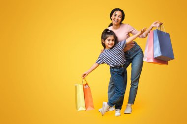 Annemle alışverişe gidiyorum. Mutlu Orta Doğulu anne ve küçük kızı bir sürü parlak alışveriş torbası taşıyor ve kameraya gülümsüyor, sarı stüdyo arka planında alışveriş yapan aile pozu veriyor, boşluğu kopyalıyor.