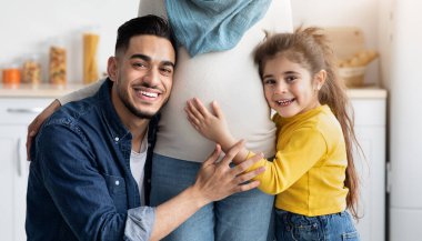 Mutlu Arap baba ve küçük kızı, tesettürlü Müslüman Anne 'nin şefkatli karnına dokunuyor, neşeli bir Orta Doğulu aile bebek bekliyor, mutfak içinde birlikte poz veriyorlar,