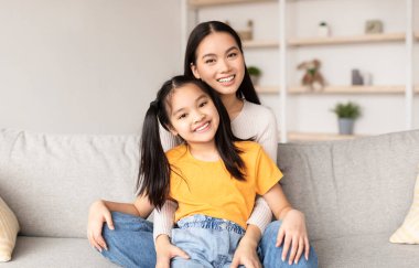 Glad smiling young korean female hugging teen girl sitting on sofa in living room interior. Mom and daughter, relationship, time together and happy family at home during COVID-19 outbreak, copy space