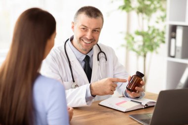 Ziyaretçi Doktor. Gülümseyen kıdemli doktor kadın hastalara haplar gösteriyor, plastik şişeyi işaret ediyor, reçete yazıyor.