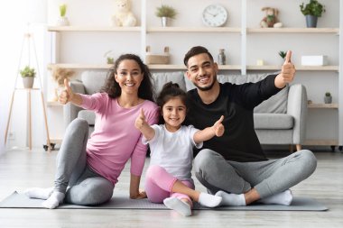 Sportif şirin bir aile, orta doğulu bir baba, anne ve küçük kızı spor kıyafetleriyle evde birlikte egzersiz yapıyor yoga minderinde oturuyor, gülümsüyor ve baş parmaklarını kaldırıyor, oturma odasının içini gösteriyor, fotokopi çekiyor.