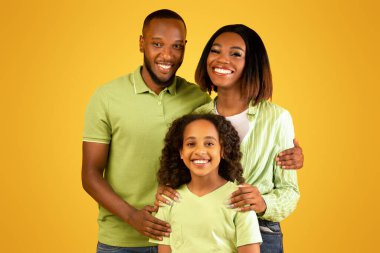 Aile sevgisi kavramı. Sarı arka planda izole edilmiş siyah aile portresi ve kızının kameraya sarılıp gülümsemesi. Annem ve babam stüdyoda çocukla poz veriyorlar.