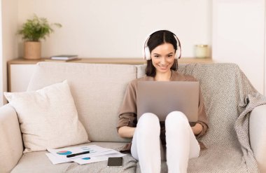 En sevdiğin şarkı. Dizüstü bilgisayarda çalışan neşeli bir serbest yazar kanepede oturuyor ve kulaklıkla müzik dinliyor.