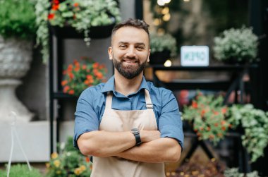 Stüdyo, kafe, dükkan sahibinin olumlu duyguları. Neşeli, kendine güvenen, önlüklü, göğsünde çapraz kolları olan sakallı yakışıklı bir adam. Çiçekçi arka planında, bitkilerle, kopyalama alanında.