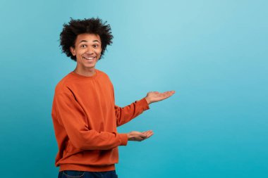 Kıvırcık saçlı neşeli, genç bir Afrikalı Amerikalı ellerini uzatıyor, parlak mavi bir arka planda boş bir alana dikkat çekiyor, terfi ya da duyurular için mükemmel..