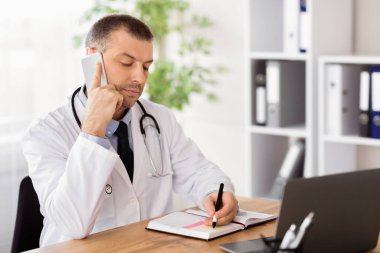 Yüksek Kalite Hizmet Konsepti. Kendine güvenen erkek terapistin portresi. Akıllı telefonuyla konuşuyor ve defterine yazıyor. Randevu hakkında notlar alıyor, müşteriye danışıyor. Uzak İletişim