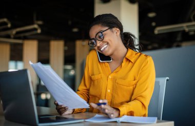 Corporate communication. Happy black female manager working with papers and talking on mobile phone, reading financial documents while sitting at workplace in office