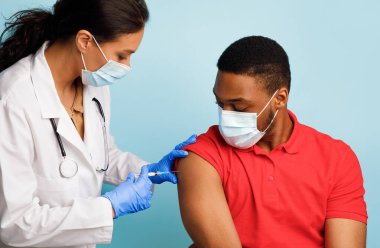 Coronavirus aşısı. Siyah Adam, Covid-19 'a karşı aşı oluyor, Doktor, Mavi Stüdyo' nun arka planına kaslar arası enjeksiyon yapan, maske takan,