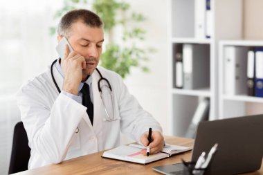 Genel Uygulayıcı Konsepti. Hastanın cep telefonuyla konuşan orta yaşlı doktor portresi. Günlüğüne not alıyor, randevu alıyor, danışmanlık yapıyor. Teletıp