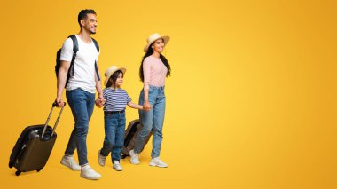 Üç çocuklu neşeli ortadoğu ailesi birlikte yaz tatiline çıkıyorlar, neşeli Arap ebeveynler ve küçük kızları sarı stüdyo arka planında bavullarıyla yürüyorlar, panorama