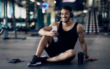 Egzersiz Uygulaması. Spor salonundaki eğitimden sonra Smartphone kullanan mutlu Arap erkek sporcu, gülümseyen Orta Doğulu adam cep telefonuyla yerde dinleniyor, internete bakıyor ya da favori müziği dinliyor, Space 'i kopyalıyor.