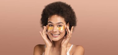 Afro-Amerikalı kadın, yanaklarında altın bir maskeyle özgüvenini kucaklıyor, gülümsüyor ve rahat, ıssız bir yerde şımartıcı rutininin tadını çıkarıyor..