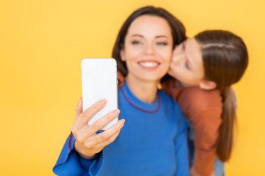 Bir kadın küçük kızıyla selfie çeker, o da neşe içinde yanağını öper. İkisi de canlı sarı bir zemine gülümsüyor birbirlerine olan yakınlıklarını ve sevgilerini gösteriyorlar..
