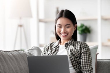 Memnuniyetle Asyalı bayan evde yeni dizüstü bilgisayar kullanıyor, koltukta oturuyor, e-posta yazıyor ya da arkadaşlarıyla sohbet ediyor, kopyalama alanı. İnternetten güzel bir film izleyen mutlu genç bir kadın. Oturma odası.