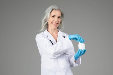 A middle aged woman in a lab coat stands confidently, holding a medical sample. She smiles warmly while wearing blue gloves, highlighting her professionalism in healthcare. clipart