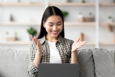 Mutlu Asyalı kadın, evinde tek başına otururken kız arkadaşlarıyla online parti veriyor rahat bir oturma odasında koltukta oturuyor, modern dizüstü bilgisayar kullanıyor, defter ekranına bakıyor ve gülümsüyor.