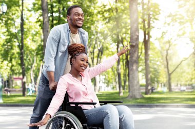 Uzun boylu, neşeli siyahi adam ve tekerlekli sandalyedeki engelli kız arkadaşı şehir parkında yürürken eğleniyorlar. Mutlu, yoksul aile, dışarıda eğleniyor.