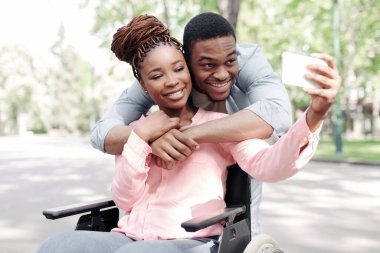 Tekerlekli sandalyedeki sevgilisine sarılan, parkta cep telefonuyla selfie çeken mutlu genç bir zenci. Afro-Amerikan çiftleri seviyorlar. Dışarıda yürürken fotoğraf çekiyorlar.