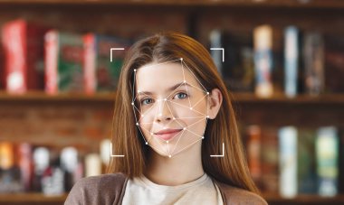 Yüz tanıma teknolojisi kitaplarla çevrili bir kütüphanede özelliklerini incelerken genç bir kadın gülümsüyor. Yüzüne odaklanarak, gelişmiş kimlik teknolojisinin kullanımda olduğunu vurguluyor..