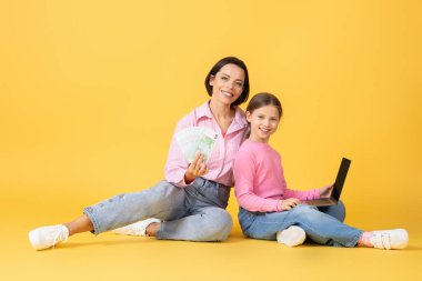 Bir anne ve küçük kızı parlak sarı bir arka planda oturup bir bağ kuruyorlar. Annesi elinde para tutarken kız bilgisayara sarılıyor, neşeyle gülümsüyor..