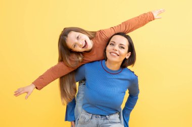 Bir anne hem gülümseyen hem de mutluluk saçan kızını neşeyle kaldırır. Parlak sarı arka plan bağlarını güçlendiriyor, birbirlerine olan sevgilerini ve sevgilerini gösteriyor..