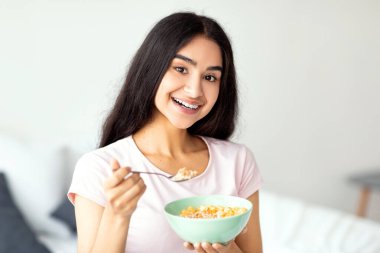 Lezzetli mısır gevreğini sütle yiyen, yatak odasında besleyici mısır gevreğinin tadını çıkaran hoş Hintli kadın. Genç Hindu kadın dengeli kilo verme diyeti yapıyor, sağlıklı yemek yiyor.