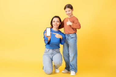 Bir anne ve küçük kızı, parlak sarı bir arka plana karşı mutlu bir şekilde poz veriyorlar, bir cep telefonunu işaret ederek. Yakın bağları sevgiyi ve sevgiyi yansıtır..