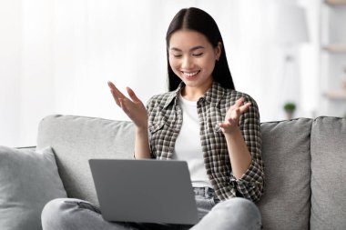 Güzel Asyalı bir bayan kanepeye oturmuş, neşeli bir şekilde el kol hareketi yapıyor, gülümsüyor COVID-19 kilitlenirken arkadaşlarıyla online parti yapıyor, mekanı kopyalıyor.