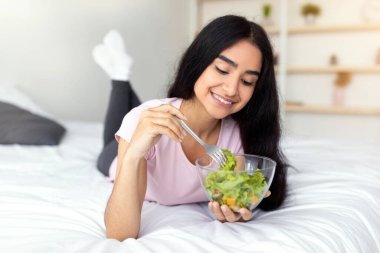 Zayıflatıcı diyet konsepti. Evdeki rahat yatakta lezzetli sebze salatası yiyen güzel Hintli bayan. Çekici Hindu kadın sağlıklı bir yaşam sürüyor, kilo veriyor, vücuduna bakıyor, evde yaşıyor.