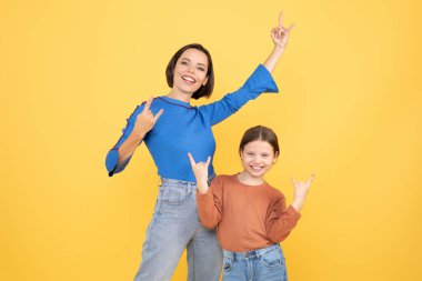 Bir anne ve küçük kızı, parlak sarı bir arka plana karşı eğlenceli ve sevgi dolu bir anı paylaşıyorlar. Neşeli pozlar verirler, aralarındaki bağı ve mutluluğu gösterirler..