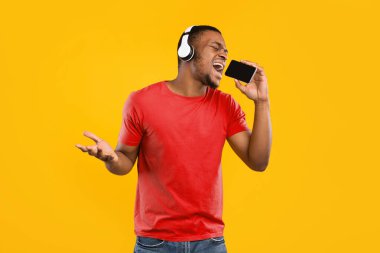 Mobil Karaoke Uygulaması. Sarı Stüdyo Arkaplan 'da mikrofon gibi kablosuz kulaklık takan Afro-Amerikan bir adam akıllı telefon tutarken eğleniyor. Çevrimiçi Müzik Uygulaması