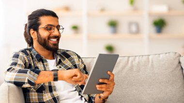 Rahat genç Hintli adam evde dijital tabletle takılıyor, kanepede oturuyor, fotokopi çekiyor. Mutlu adam internetten alışveriş yapıyor, modern ped kullanıyor ve gülümsüyor.