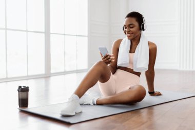 Gülümseyen Afro-Amerikan kadın yoga minderinde oturuyor, antreman arasında akıllı telefon kullanıyor, kablosuz kulaklık takıyor, spor sutyen takıyor ve beyaz havlu takıyor. Favori çalma listesini dinleyen bayan