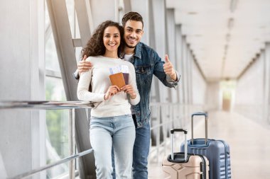 Seyahat Reklamları. Havaalanında mutlu genç Arap çift pozu, terminalde bekleyen neşeli çift, biletleri elinde tutan kadın ve kamerada başparmağını gösteren adam,