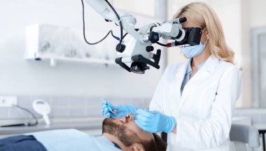 Modern mikro dişçilikte yenilikçi aletler. Yeni klinikte diş mikroskobuna bakan dişi dişçi, erkek hasta, yan görüş, kopyalama alanı için tedavi yaparken yenilikçi araçlar kullanıyor.