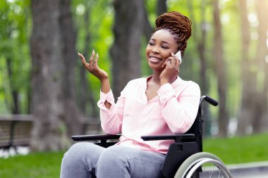 Tekerlekli sandalyedeki neşeli siyahi kadın şehir parkında arkadaşıyla telefon görüşmesi yapıyor. Engelli Afro-Amerikan kadın akıllı telefonuyla birileriyle konuşuyor, dışarıda iletişiminin tadını çıkarıyor.