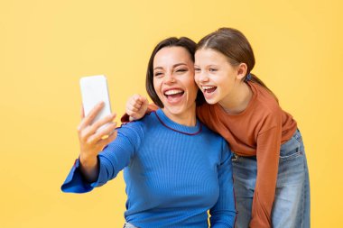 Bir anne ve kızı, parlak sarı bir arka planda selfie çekerken geniş bir gülümseme sergiliyorlar. Kahkahalar bir neşe anı yakalar ve birbirlerine bağlanırlar, sevgilerini ve sevgilerini gösterirler..