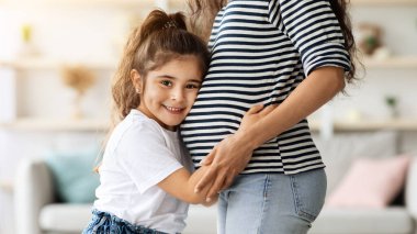 Sevimli küçük kız hamile annesine sarılıyor ve kameraya gülümsüyor, orta doğulu çocuk annesine bağlanıyor, ablası olmak için sabırsızlanıyor, yakın plan portresi