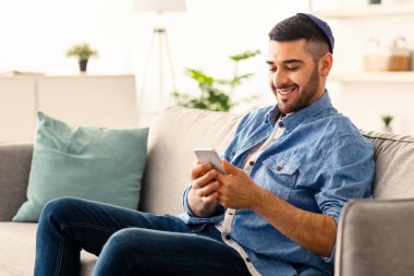 Mobil İletişim Konsepti. Evindeki rahat koltukta oturan modern akıllı telefon cihazını kullanan mutlu genç Yahudi portresi. SMS yazan neşeli adam, sosyal ağda geziniyor.