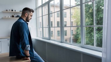 Kişisel sorunları düşünmek, yalnızlık, dram ve depresyon, covid-19. Mutsuz genç Avrupalı yakışıklı adam oturma odasının iç tarafındaki büyük pencerenin önünde duruyor.