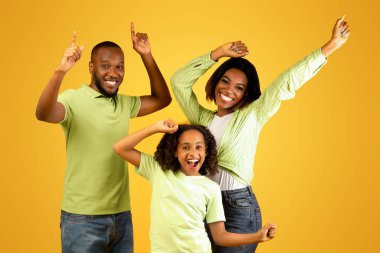 Üç kişilik heyecanlı zenci aile dans ediyor ve oynaşıyor, sarı stüdyo arka planında favori müziğin tadını çıkarıyorlar. Afro-Amerikalı adam boş alanı işaret ediyor.