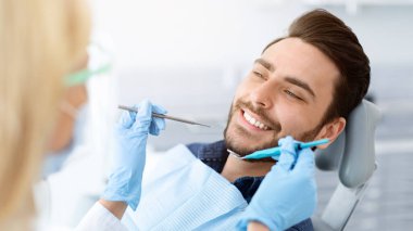 Kadın dişçi, maske takan, hasta, neşeli, dişçi aletleri tutan, lastik eldiven giyen genç adam için tedavi gören sarışın bir kadın. Stomatoloji, modern diş sağlığı kliniği kavramı