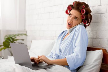 Kafası karışmış ev hanımı cep telefonuyla konuşuyor dizüstü bilgisayar kullanıyor evde, modern yatak odasında yatağında çalışıyor. Mobil İletişim, Mesafe ve Serbest Çalışma. Yan Görünüm