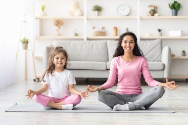 Spor giyim kuşamında rahat bir anne ve küçük kızı nilüfer kumaşında oturmuş gülümsüyor, evde birlikte meditasyon yapıyor, fotokopi çekiyor. Sağlıklı aile, spor yaşam tarzı konsepti