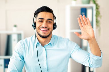 Uzaktan iletişim konsepti. Kulaklıklı mutlu bir Arap ofiste kameraya el sallıyor. Genç adamın iş görüşmesi ya da internet konferansı için bilgisayar kullanırken görüntüsü.