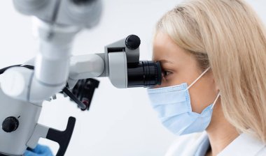 Çalışan dişçi modern diş mikroskobuna bakarak, tedavi için yenilikçi aletler kullanıyor. Sarışın kadın doktor gıcır gıcır dişçi kliniğinde tedavi için en yeni ekipmanları kullanıyor.