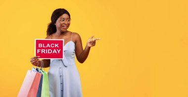 Elinde alışveriş torbaları olan çekici genç bir zenci kadın. Elinde Kara Cuma tabelası var ve boş alanı işaret ediyor. Turuncu stüdyo arka planı. Afro-Amerikan bayanın indirim günü.