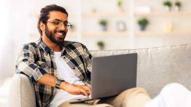 Gözlüklü mutlu Hintli adam evde çalışıyor, modern not defteri kullanıyor, rahat oturma odasında kanepede oturuyor, fotokopi çekiyor. Gülümseyen Orta Doğulu adam serbest programcı ya da kadınlarla sohbet ediyor.