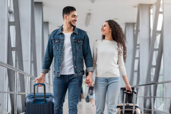Portrait Of Romantic Middle Eastern Spouses Walking With Suitcases At Airport Hall, Happy Young Arab Man And Woman Going To Departure Gate With Luggage And Smiling To Each Other, Free Space